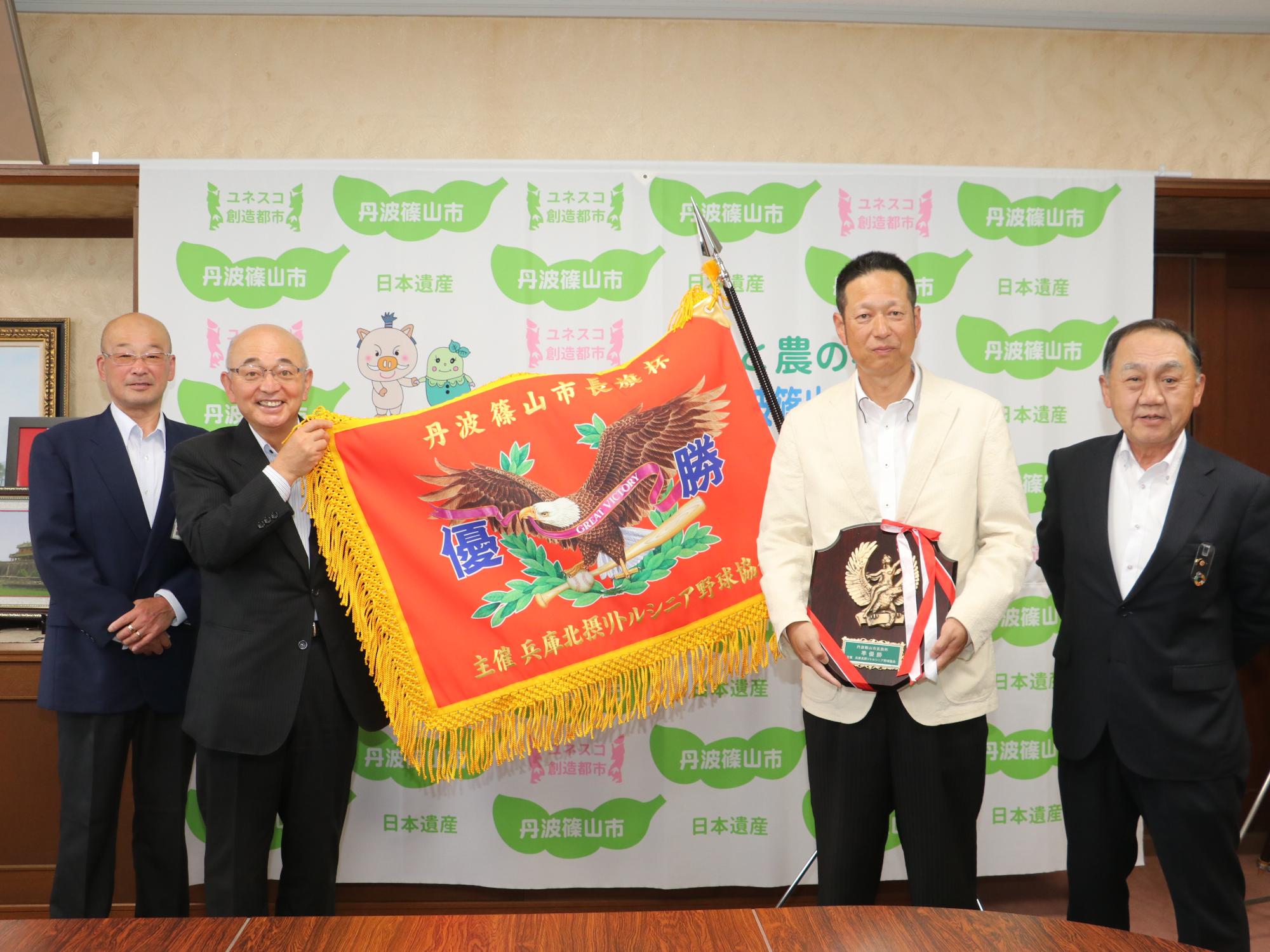 市長杯の優勝旗を市長が手に持って広げていて、盾を持った男性、両隣にスーツを着た男性が立っている