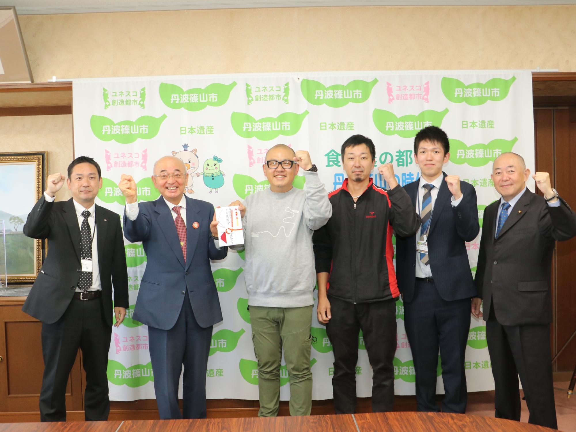 奨励金を手にガッツポーズする中西さんと市長、関係者の皆さん
