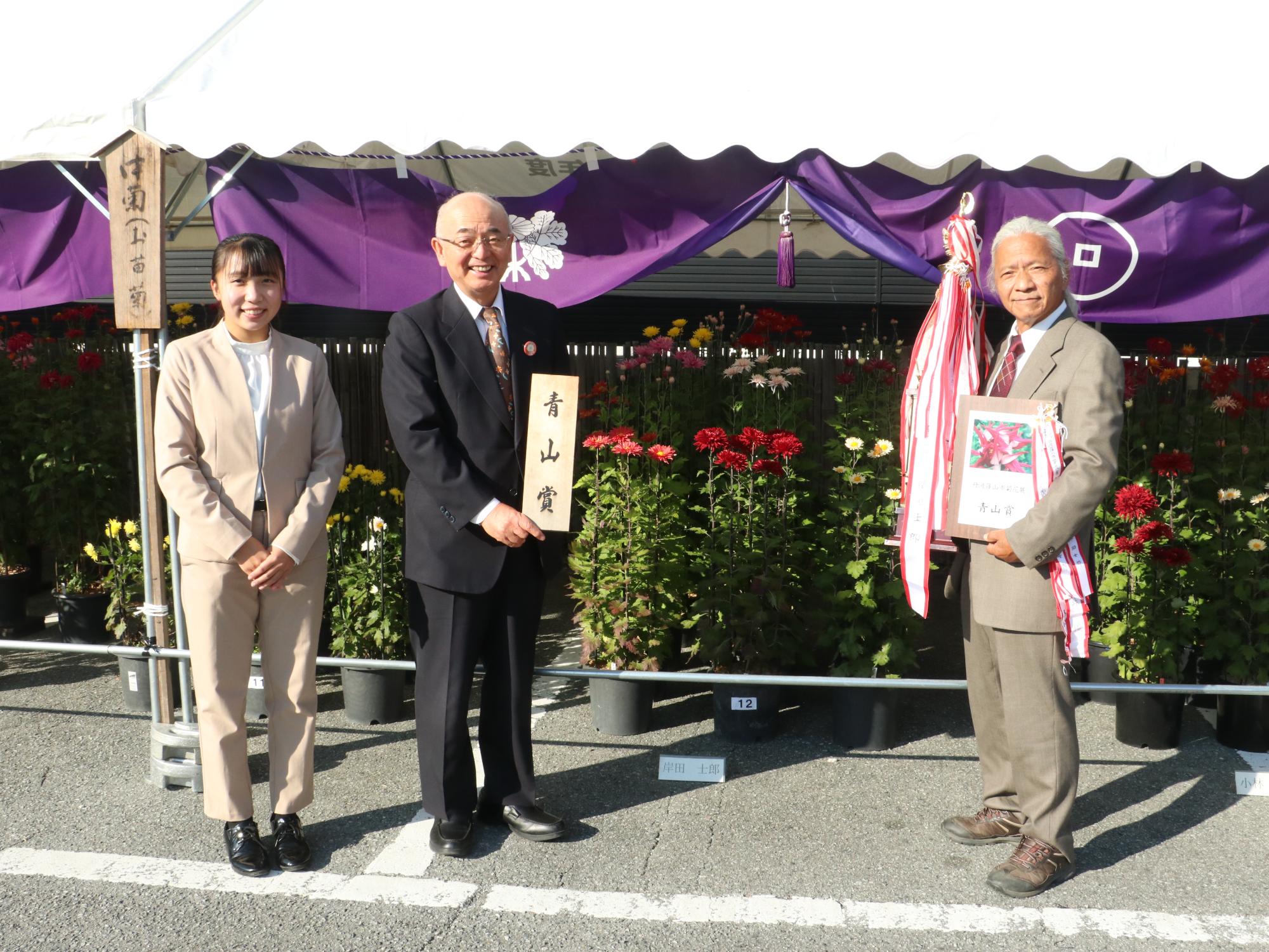 菊花展で、市長が青山賞のプレートを持ち、右隣で受賞者が青山賞の盾とトロフィーを抱えている、左隣にほかの受賞者が立っている