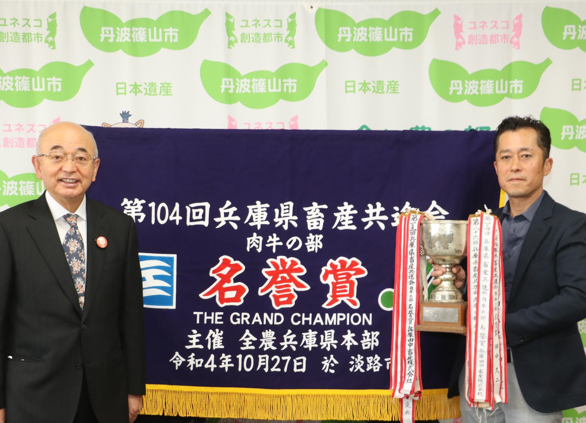 名誉賞の横断幕の前で田中さんがトロフィーを持ち、市長と記念撮影