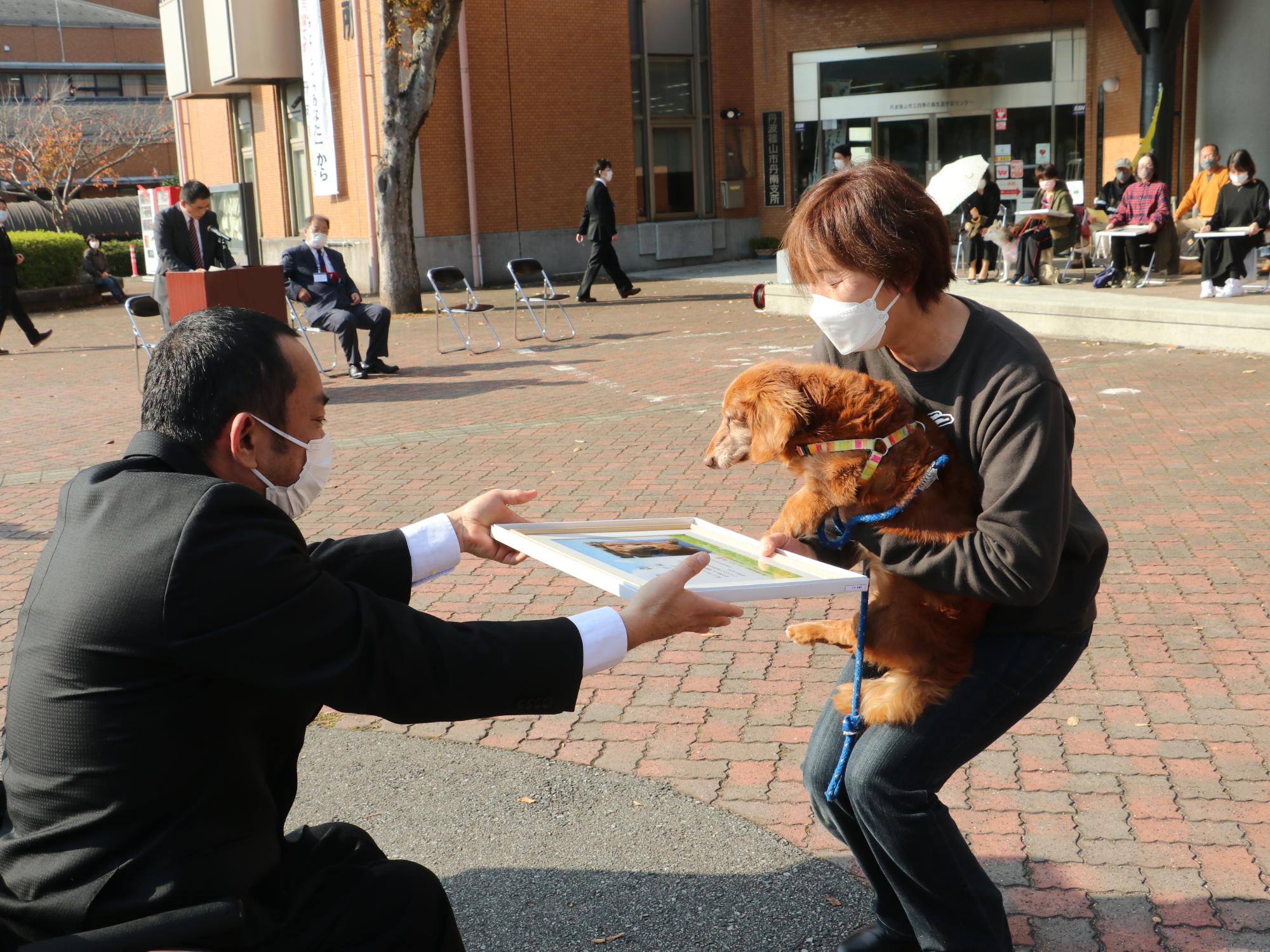 犬を抱っこしている飼い主に表彰状を手渡している