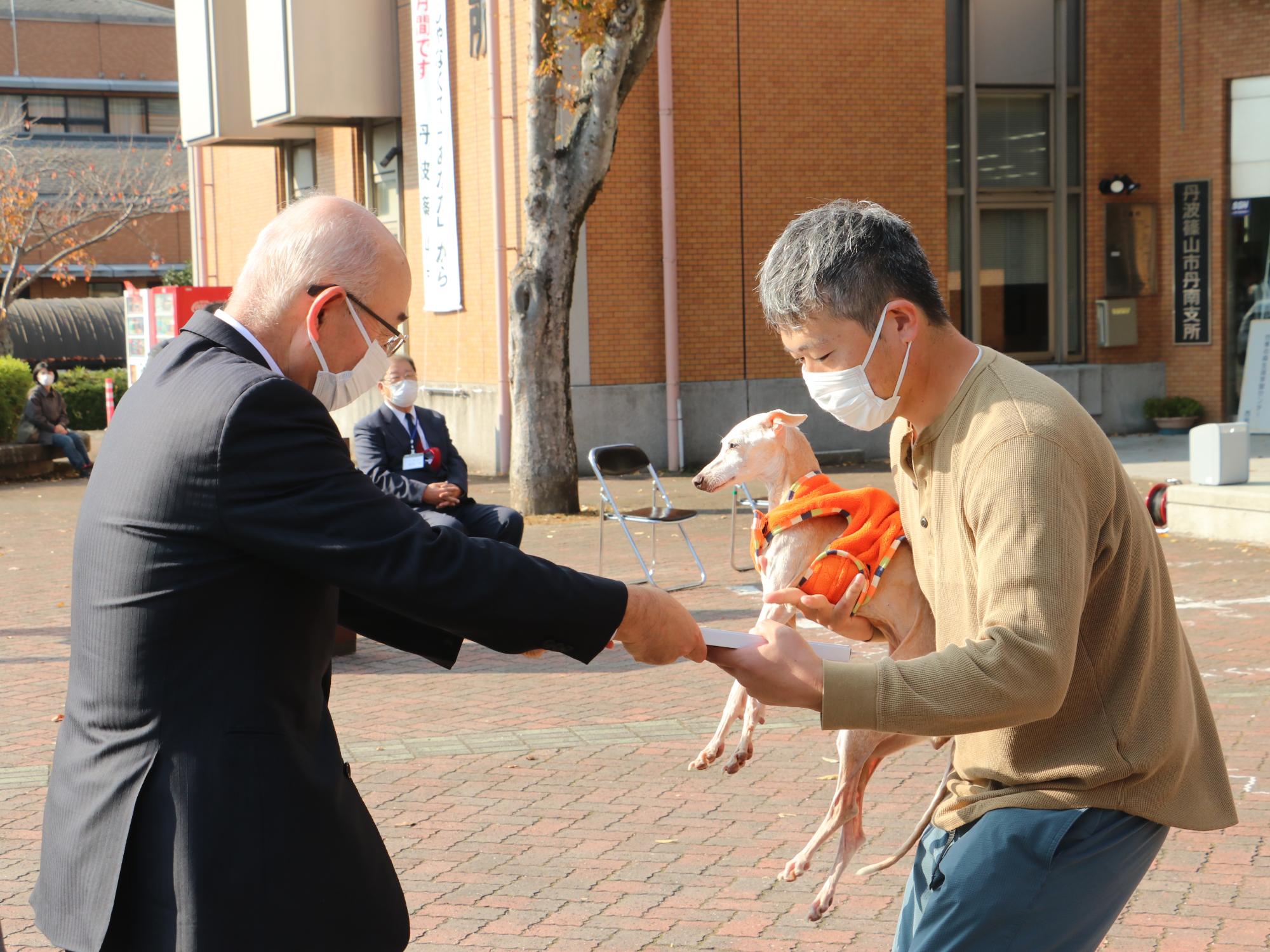 犬を抱っこした飼い主に表彰状を手渡している