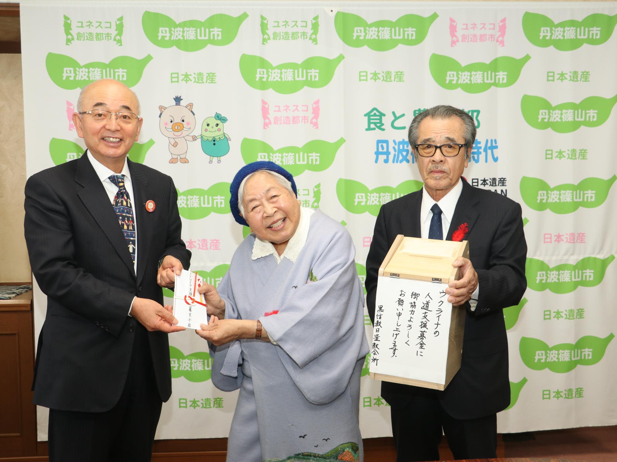 女性が募金の目録を市長に手渡している。その隣に、募金箱を持った男性が立っている