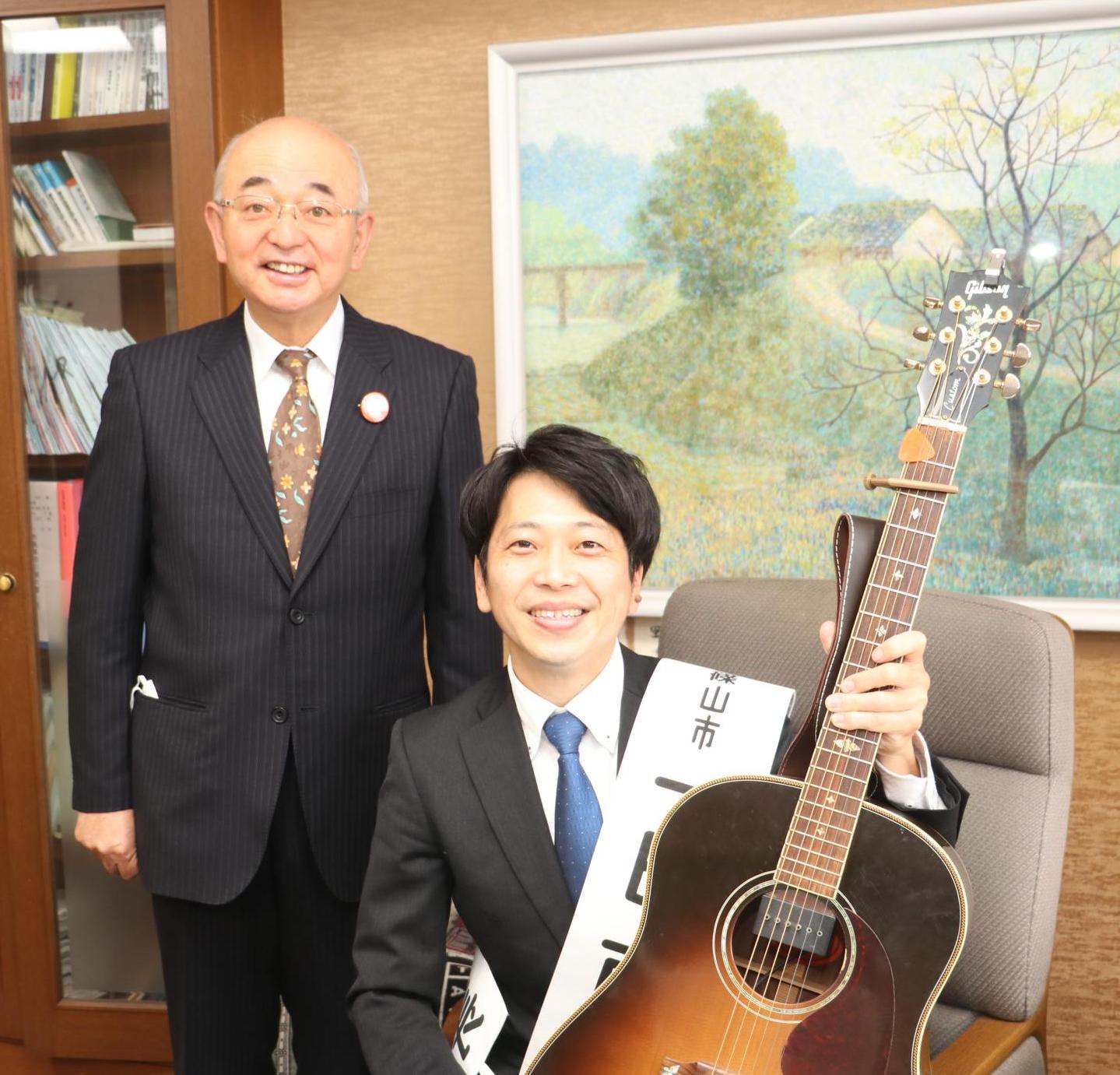 1日市長の雄介さんがギターを持ち市長の椅子に座り、その隣に酒井市長が立っている