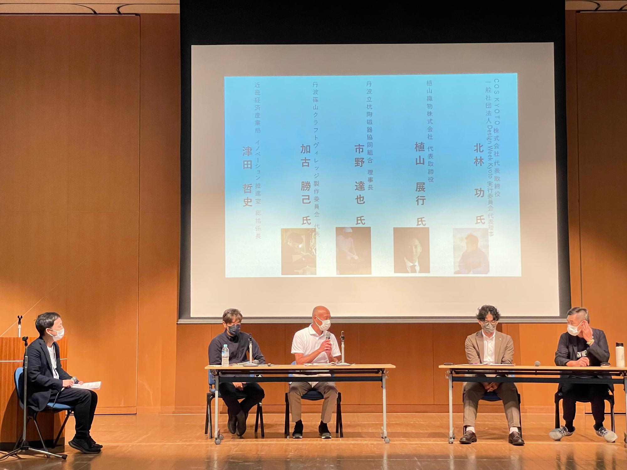お名前が紹介されたスライドの前に、パネルディスカッションをされる4名の男性が座って話をされている