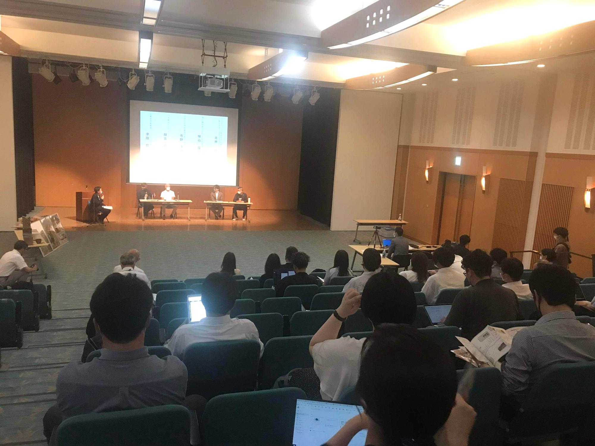 会場の後方から、フォーラムの様子を撮影している。舞台にコーディネーター1名、パネルディスカッションをされる4名の方が座っている。