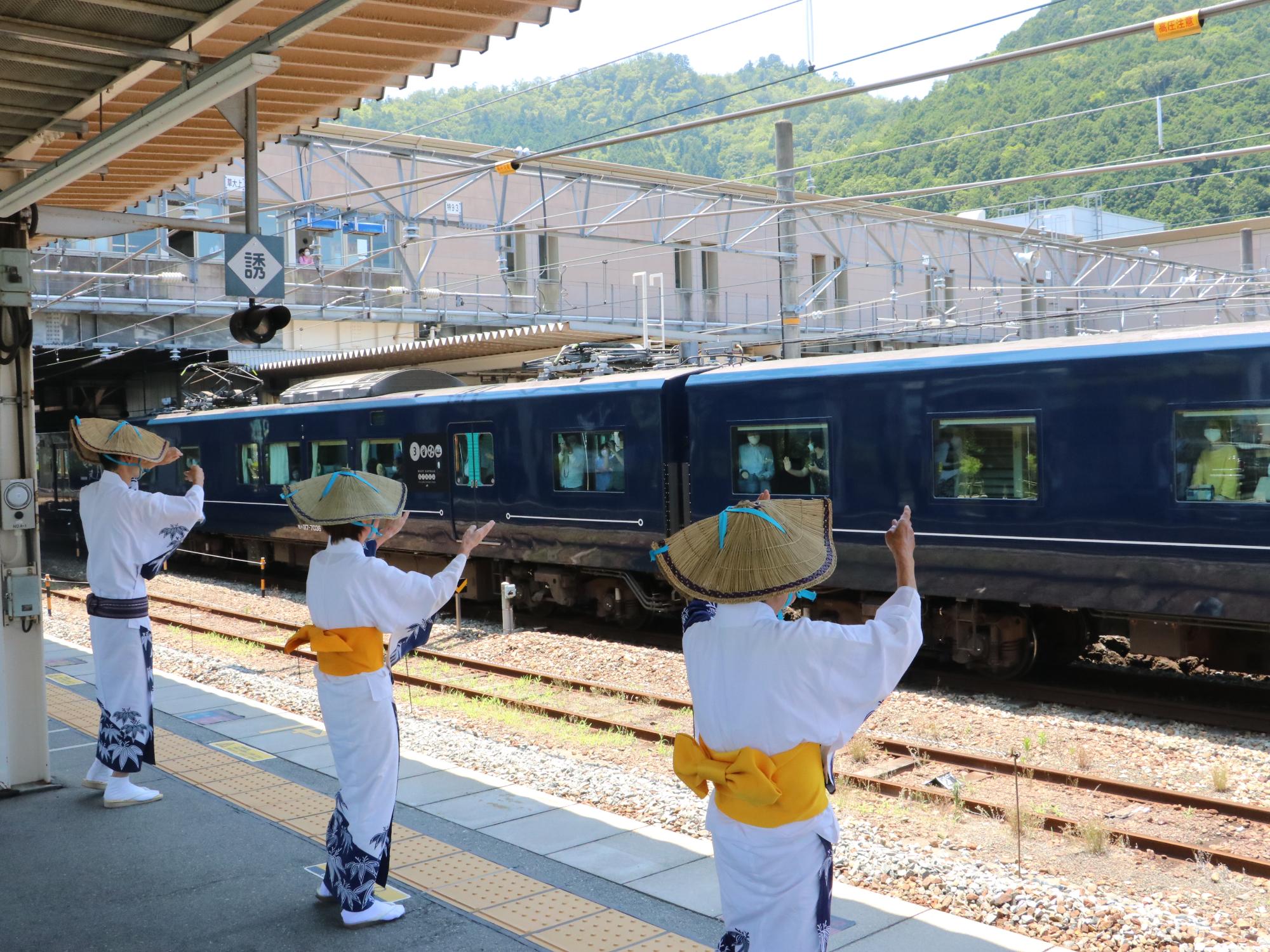 駅のホームで、笹浴衣を着て編み笠をかぶった方数名がデカンショ踊りを踊っている。その様子を、向いのホームに停車している観光列車「銀河号」の乗客が見ている