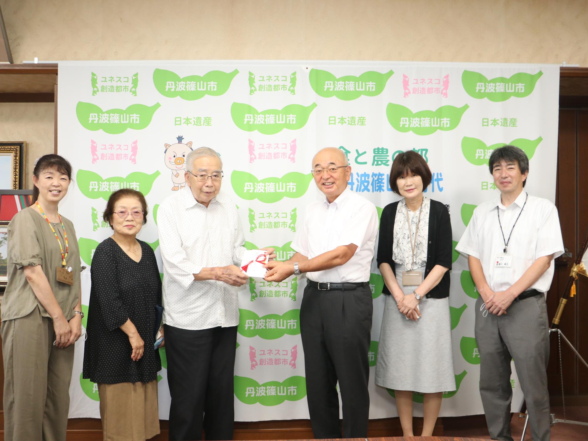 元助役がが寄附の目録を市長に手渡している。隣に奥様と現役時代に元助役に育てられた職員数名が並んでいる。