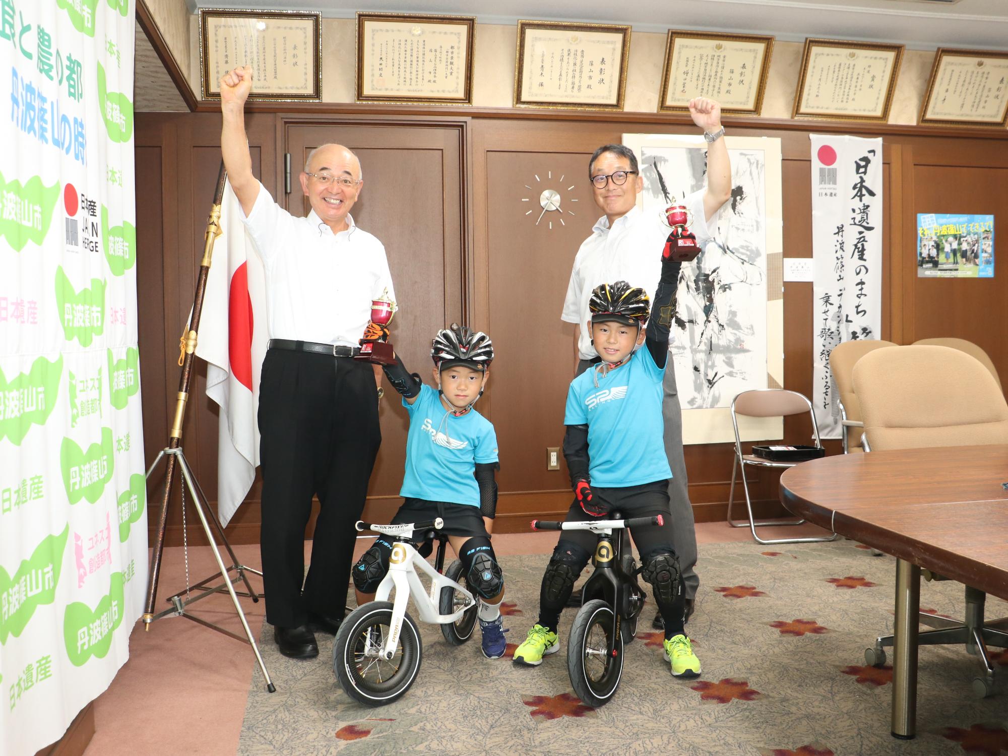 男の子二人がランバイクに乗っていて、それぞれカップを持った手をあげている。後ろに市長と教育長が片手をあげて立っている。