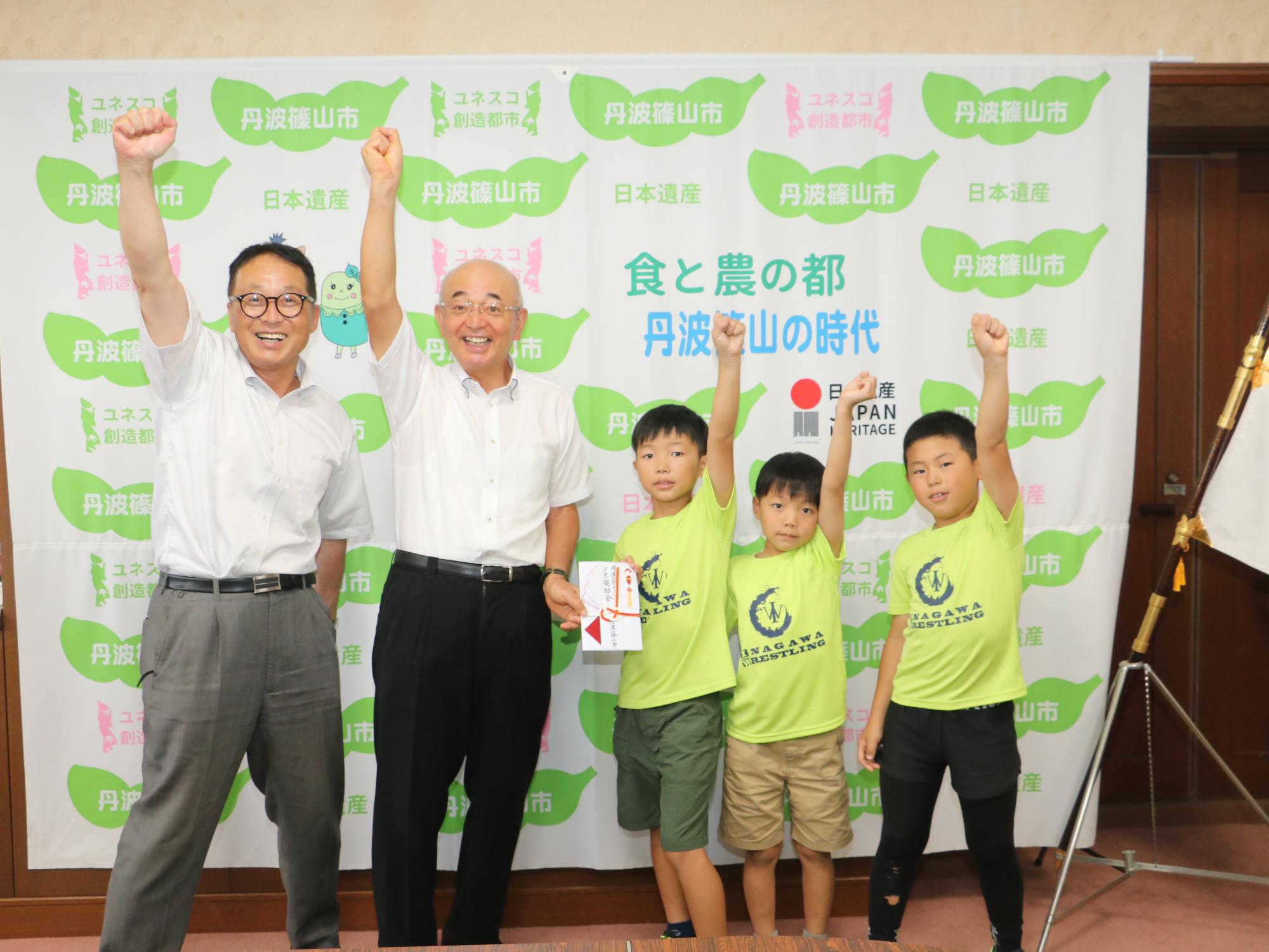 一人の男の子が奨励金の目録を持っていて、片手をあげている。その両隣に男の子二人と市長、教育長が片手をあげて立っている