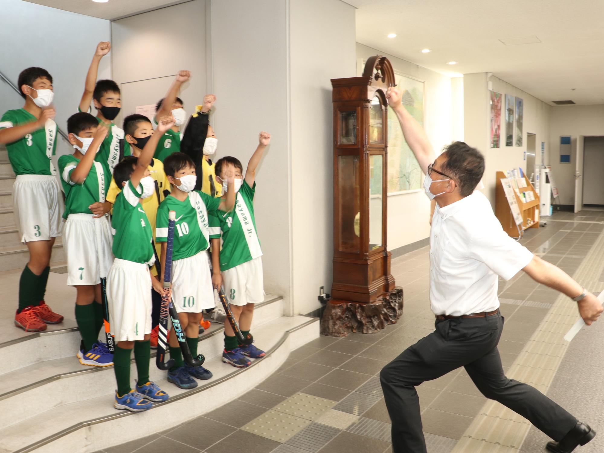 階段に、ホッケーのユニフォームを着た男の子たちが並んで片手をつきあげていて、前で教育長も片手をつきあげている。