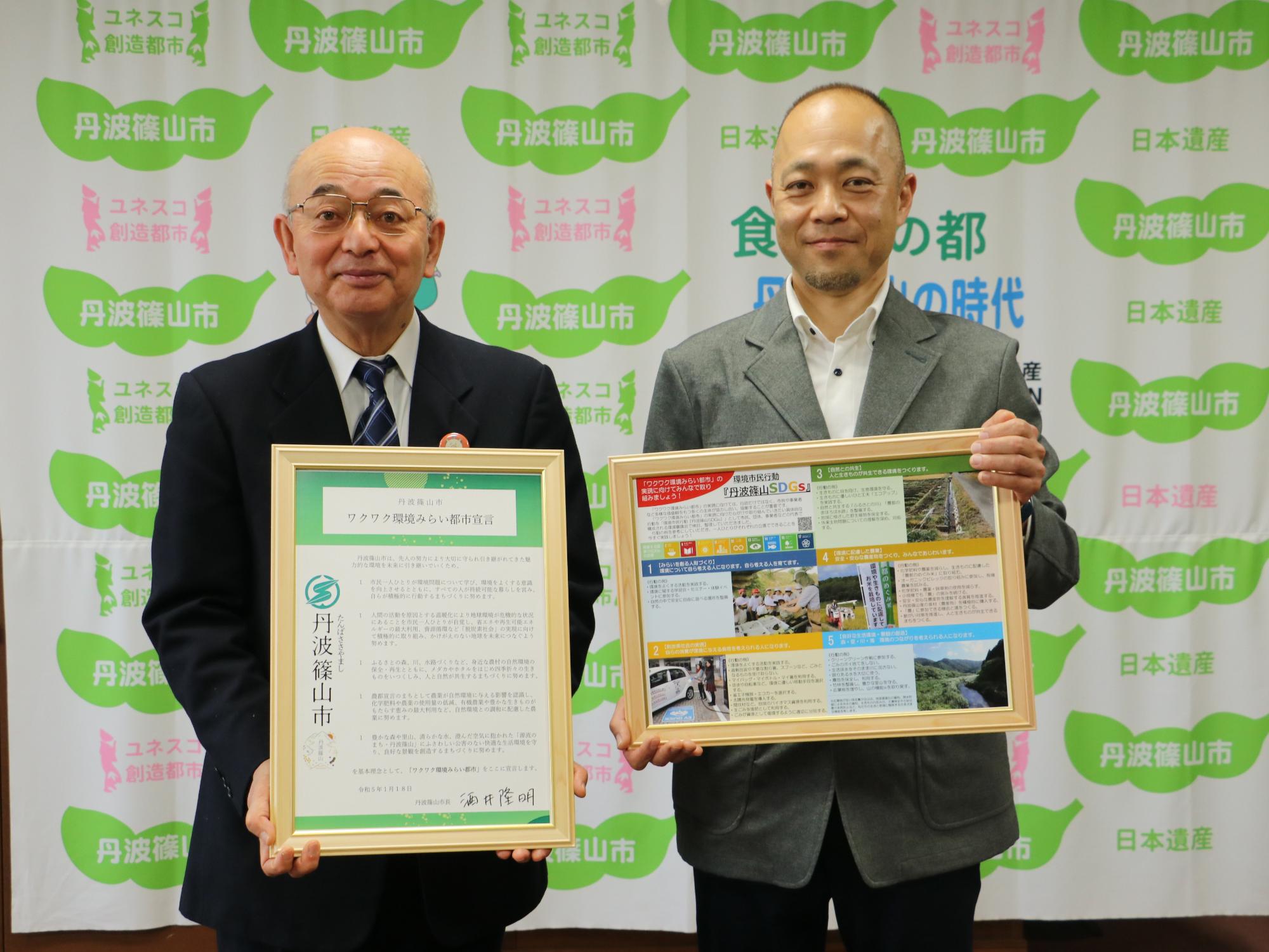 市長が宣言を手に持ち、審議会会長がSDCSの取り組みのパネルを持っている