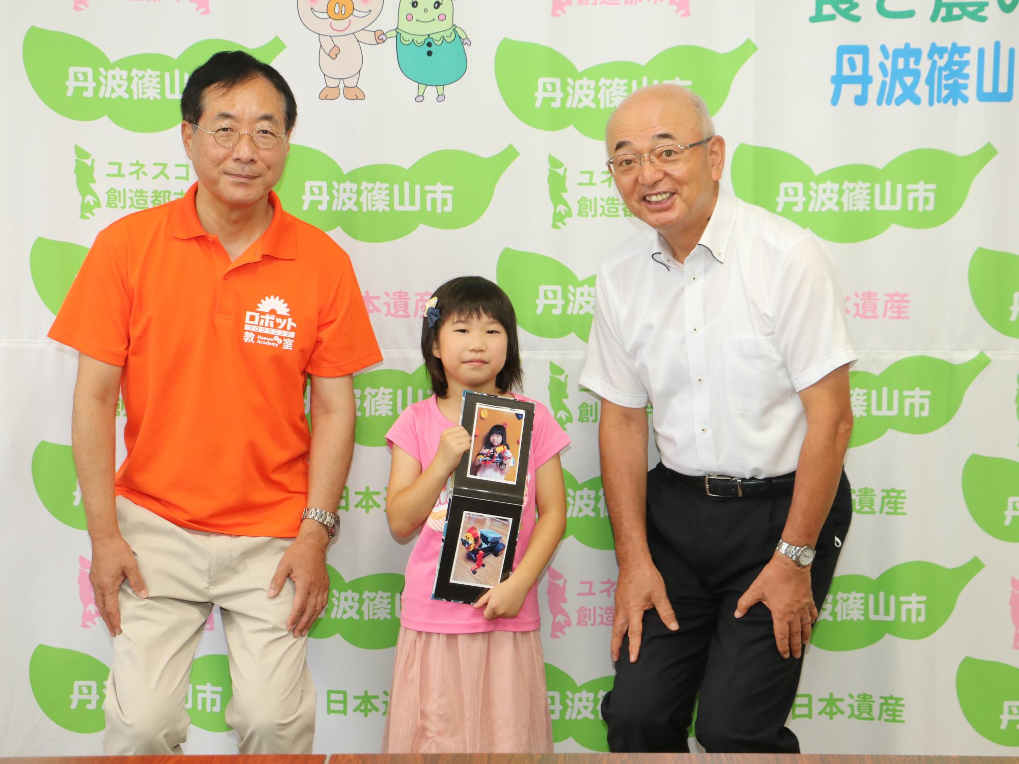 作ったロボットの写真を持った女の子を真ん中に、市長と、先生が立っている