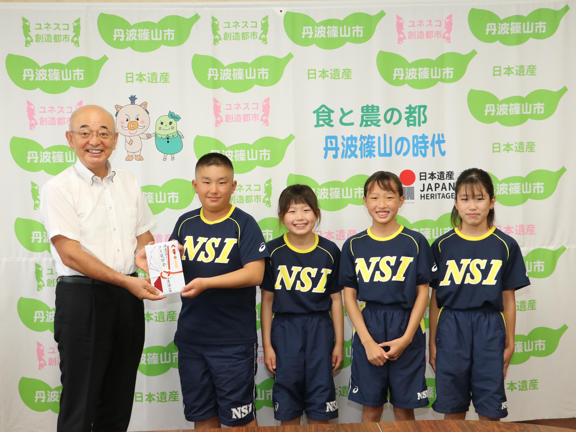 市長が水泳クラブのユニフォームを着た男の子に奨励金を渡している。男の子の隣には女の子が3人立っている