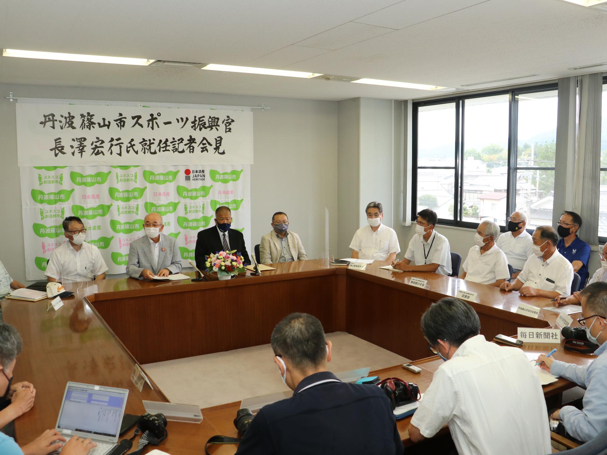 会議室奥にタペストリーと手書きで書かれた記者会見の横断幕がある。市長、教育長、長沢先生が前方に座っていて関係者や記者が周りに座っている