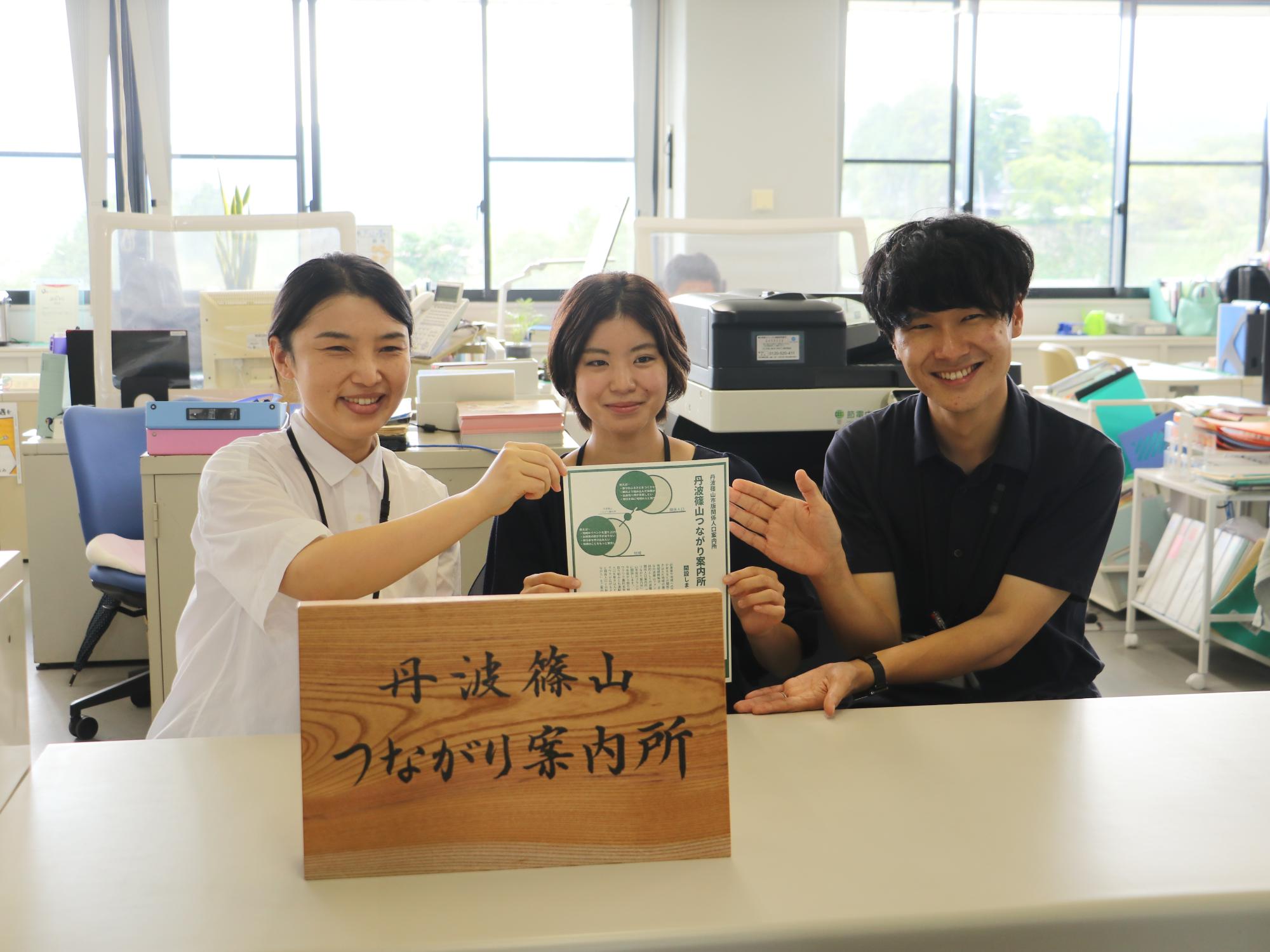 カウンターに新しくできた案内所の看板を置き、後ろで若手職員3人がチラシを持っている