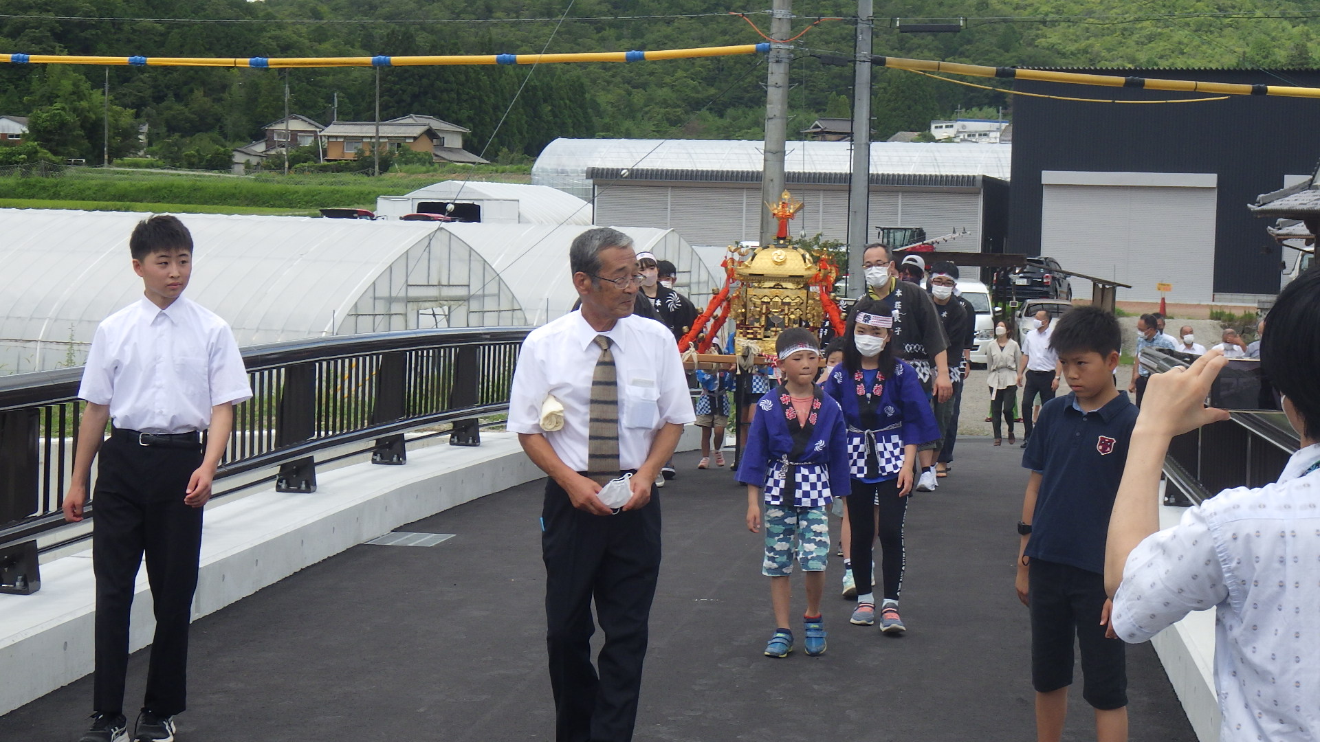橋の渡り初めで、ハッピを着たこどもや大人が神輿の周りに集まっており、先頭に地元の役員や子どもたちがいる。