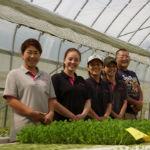 ポロシャツ姿の呉田さんと従業員4名がハウスの水耕栽培野菜の前で笑顔で横一列に並んだ写真