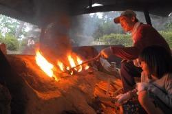おじいさんと子供たちが土の山から出ている火を薪や棒で火力調整している写真