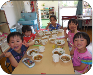 机を囲んで笑顔でご飯を楽しむ園児たちの写真