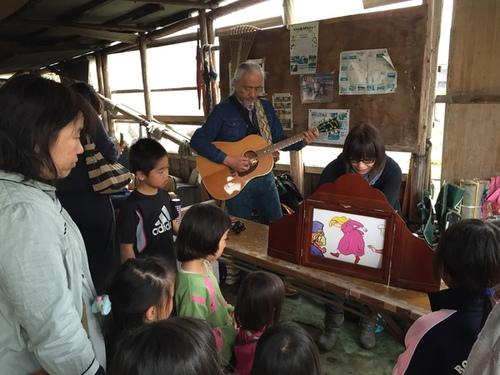 ゆるフリマで紙芝居をする女性と紙芝居に合わせてギターを演奏する男性と見つめる女性と子供達の写真