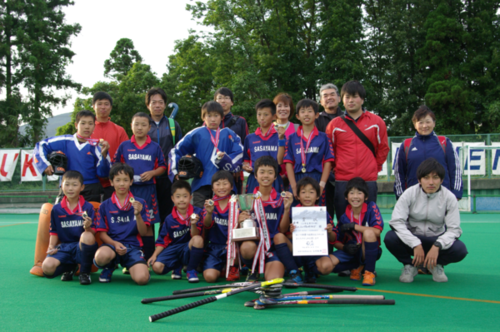緑のコートでトロフィーと賞状を持って笑顔を見せる篠山ジュニアホッケークラブ小学男子チームの選手達の写真