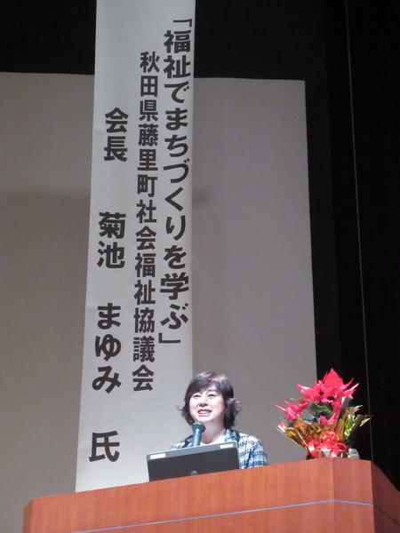 ステージ上に設置された「福祉でまちづくりを学ぶ」と書かれた垂れ幕の前で、マイクの前で講演を行う菊池まゆみさんの写真