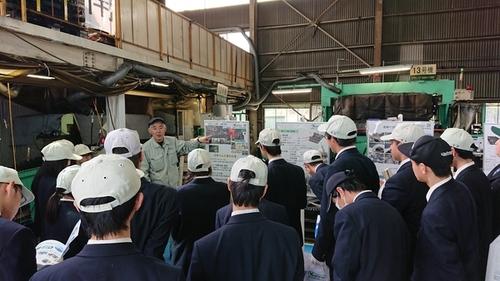吉野ゴム工業株式会社の13号機と書かれた緑色の機械のそばでパネルを指さし説明を行う職員の方と、白い帽子をかぶり説明を聞く生徒たちの写真