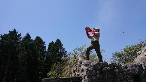 青空の広がる頂上の崖に立ち、ホラ貝で御嶽と弥十郎ヶ嶽の間で交信を行っている様子の写真