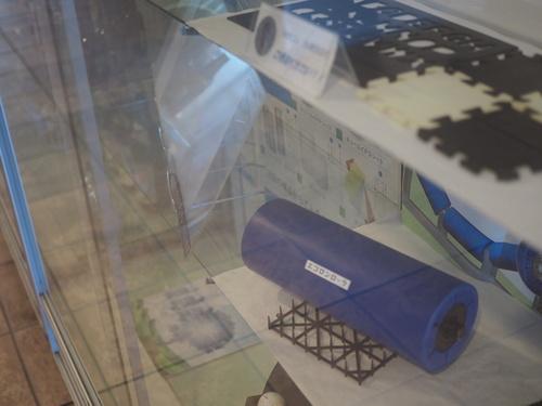 篠山市企業紹介コーナーのエコロンローラと書かれた青筒の工業部品等の展示物を映している写真