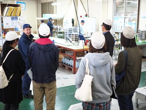 保護者の方々が白い帽子をかぶって芦森工業のホースなどの製造工場で説明を聞いている様子の写真