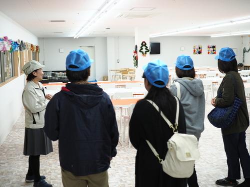 保護者の方々が帽子をかぶって岩崎電機製作所の新設工場を見学している写真