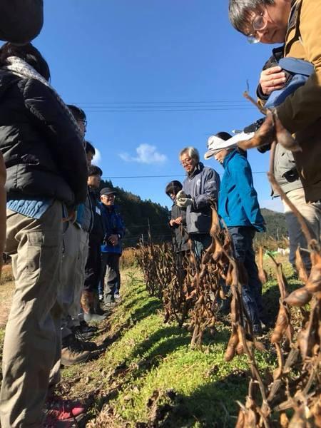 青空の下で行われた体験プログラム「川原地区での黒豆オーナーの畑見学」で実った黒豆を前にしている参加者達の写真