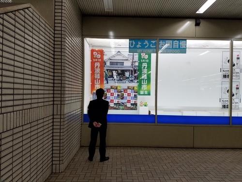 ショーウインドーの中に飾られた丹波篠山市誕生を発信するポスターを後ろに腕を組んで見る男性の後ろ姿の写真