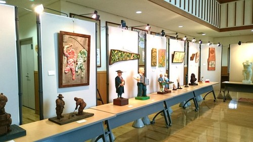 ライトが点いた展示ボードにかけられた壁掛けの彫刻作品とテーブルの上に展示された様々なモチーフの彫刻作品の写真