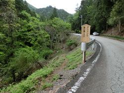 クリンソウ登山道入り口と書かれた木の立て看板がガードレール脇に立つ山あいのカーブしたアスファルト道路の写真