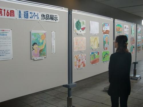 第16回『農』絵画コンクール作品展示と書かれたパネルに貼りだされた作品たちを鑑賞する1人の女性の写真