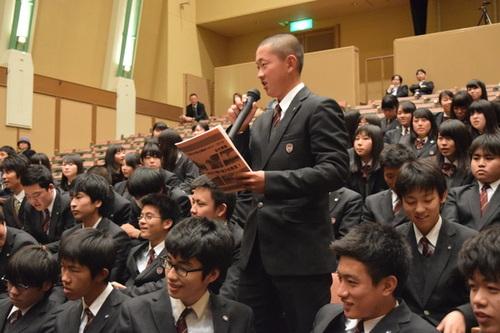 パンフレットを左手に持ち、右手にマイクを持って観客席から立って質問をする男子高校生の写真