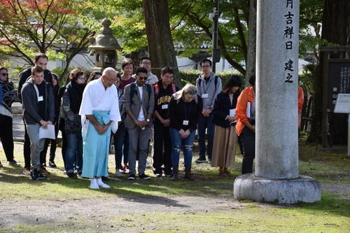 神社の参拝方法のレクチャーを受け、白衣に水色の袴を着た宮司さんと一緒にお辞儀をしている参加者の方たちの写真