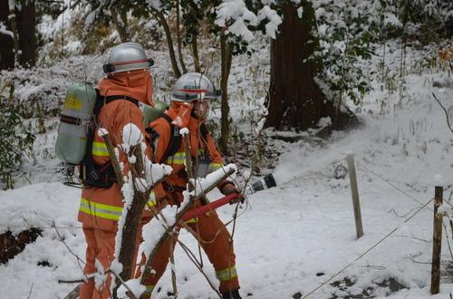 オレンジ色の作業着を着て雪山の中で放水作業をする二人の人物の写真