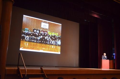 多目的ホール壇上スクリーンに剣道教室の集合写真を写して発表する堀毛孝さんの写真