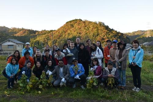 紅葉で色づいた山々を背景に2列になって収穫した枝豆を手に笑顔で写真に写るツアー参加者の方たちの写真