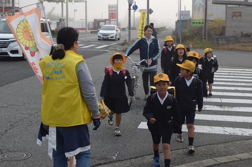見守りを兼ねて道路を横断する通学児童に挨拶をする黄色のベストを着用した地域の人・保護者・学校の先生達の写真