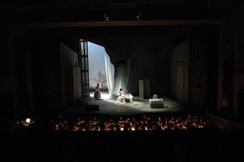 手前にオーケストラ奏者が並び、奥の舞台で窓際に立つ女性と手前に座る女性とのシーンの写真