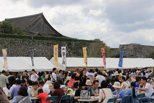 篠山城跡三の丸広場にカラフルな登り旗が立つ屋台の前で、食事を楽しむ大勢の人たちの写真