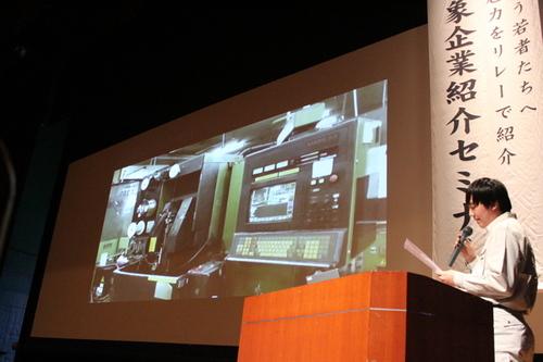 緑と黒の大きな機械が映し出されたスクリーンの横で話をする男性の写真