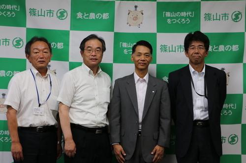 篠山市のスローガンが書かれた白と緑の幕の前に立つスーツを着た4人の男性の写真