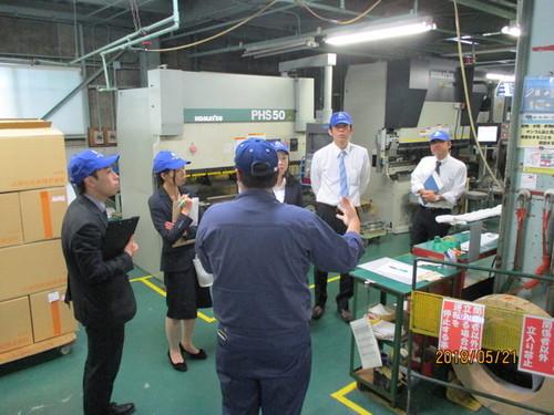伸和工業株式会社にて、大きな機械の前で青い帽子をかぶった職員の方の説明を聞く教職員の皆さんの写真