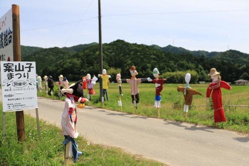 個性的な案山子がたくさん山のふもとの田んぼの端に並んで立っている様子の写真