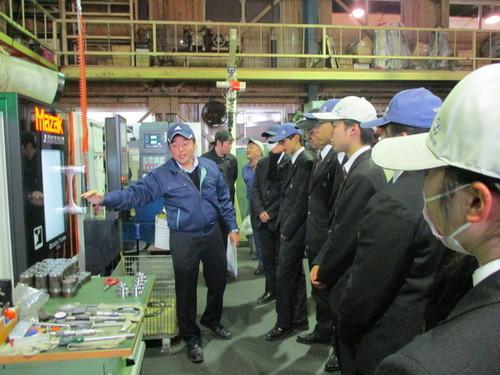 帽子を被り工場見学をする高校生たちが大型機械と工具類を前に担当者から説明を受ける写真